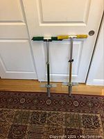 Pair of metal pogo sticks standing upright against a white door on a patterned rug. One pogo stick has green foam handles, the other yellow. Both have metal springs, foot rests, and adjustable height.