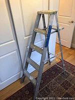 5 foot aluminum step ladder with four steps and a top platform, positioned indoors on a hardwood floor next to a white door.