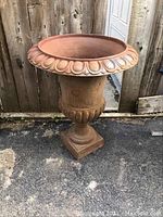 Front angled view of rust-colored cast iron urn with decorative rim and square base, placed on outdoor pavement.