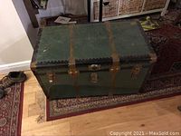 Closed green steamer trunk with brown leather straps and metal clasps on a wooden floor.