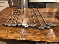 Eight right-handed Ping golf irons laid out on a wooden table, showing the full length of shafts and heads to illustrate condition and set completeness.