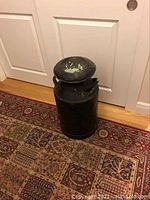 Full view of black metal milk jug on patterned carpet showing jug's cylindrical shape and lid with paint wear.