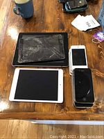 Top view of multiple Apple devices on wooden table including iPhones and iPads, one iPad with a cracked screen