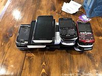Stack of many Blackberry phones in two rows, some with physical QWERTY keyboards, some touchscreen models, various colors (black, white, red).