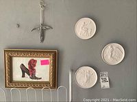 Wall grouping showing gilt framed shoe picture, three plaster Greek figure plaques, and metal angel hanging above