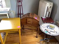 View of five small tables including yellow desk, sewing cabinet, small dark pedestal table, tile-top side table, and metal easel stand.