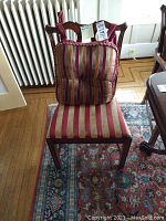 Front view of a Duncan Phyfe style dining chair with striped red and gold padded seat and a matching tufted striped cushion tied to the backrest. Chair is on a patterned carpet next to a radiator.