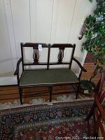 Full front view of vintage wooden bench with green fabric cushion and carved backrest design placed on a woven rug indoors.