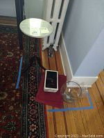 Photo showing the small round marble-topped table with tripod base, clear glass bowl with broken edge, and artificial tree with red berry decorations in white pot on a wooden floor beside a radiator and patterned rug.