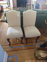 Full view of two white fabric upholstered side chairs with tall backs and dark wooden legs, placed against a wall in a room with wooden floor and fireplace in the background.