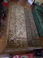 Full view of tan floral print wool area rug laid out on a wood floor near a green couch and fireplace.