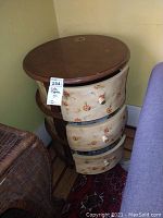 Round side table with a wooden top and three floral drawers with signs of wear and scratches, shown from an angle with all drawers partially open.