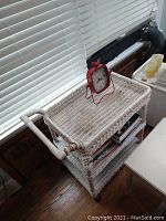 White wicker cart against window, filled with books in lower shelves and red clock on top shelf.