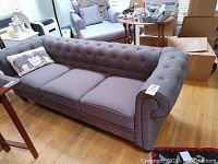 Front side view of gray tufted three-seat sofa with rolled arms and canvas upholstery, showing decorative elephant pillow on left end.