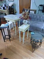 Four side tables in a room: two painted distressed wooden tables, one metal round table with glass top, and vintage folding table; wooden floor and sofa in background.