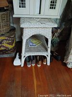 Front view of white wicker table showing woven construction, open shelf, and shoes stored beneath. Top white painted wicker surface visible.