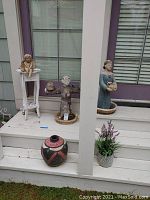 Outdoor resin figurines displayed on steps including seated angel on white stand, standing angel, bust figurine, and decorative vase on ground.