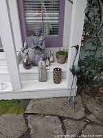 Front view of outdoor decorations on porch step including resin figurines, wooden figurine, lanterns, glass planter, and tall metal stork statue on ground.