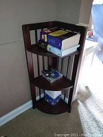 Dark wood corner folding shelf unit with three shelves, placed in a room corner, holding books and small items.