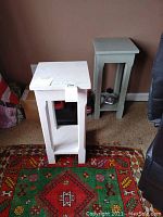 Two matching wooden lamp tables, one white and one light green, shown on a colorful patterned rug against a wall.