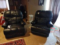 Two black leather reclining chairs side by side on carpeted floor with natural light from window, showing front and angled view of chairs including armrests and reclining mechanism lever.