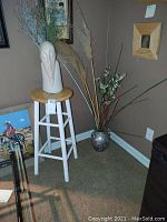 Full view of wooden stool with white sculptural vase holding dried plants and metallic decorative vase with dried botanicals on carpeted floor corner.