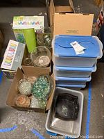 Photo showing box of 16 acrylic tumblers, two medium glass bowls and a small glass vase, several bags of decorative glass gems in various colors, and stacked clear plastic storage containers with blue lids along with two metal tins stacked inside a larger plastic container.