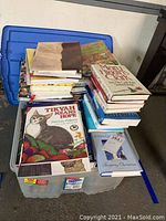 Photo of assorted books stacked on top of a large plastic storage bin, including children's books and cookbooks visible on top.