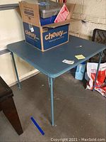 Green folding card table with metal legs, scratches, rip on top, box of scrapbooks on table