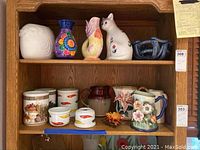 Wide view of shelves showing multiple porcelain and ceramic decorative items including vases, pitchers, mugs, cat sculpture, and blue bowl.