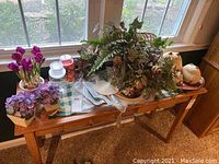 Long view of wooden table with various faux flower arrangements, decorative plates, silver platter, and glass jars scattered on top.