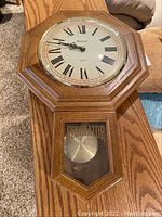 Full view of Bulova oak regulator wall clock on table showing wood frame, pendulum, and clock face with Roman numerals.
