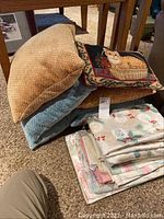Photo showing three pillows stacked under a wooden table along with folded floral and holiday-themed table linens beside them.