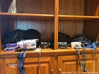 Three cameras and one camcorder displayed on wooden shelf with carrying cases behind.