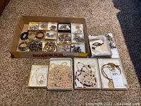Full overview of jewelry collection in boxes including necklaces, bracelets, watches, and earrings arranged in cardboard trays and boxes.