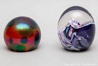 Front and side view of two glass paperweights: one iridescent with colored spots, one clear with purple swirl