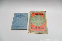 Two vintage stamp albums side by side on white surface, one blue titled 'The Improved Stamp Album' and other titled 'World's Stamp Album' with orange and green cover