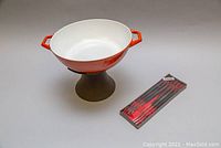 Orange enamel pan with white interior placed on cast iron stand, alongside unopened boxed fondue forks