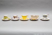 Five assorted vintage teacup and saucer sets side-by-side, showing varied floral and gold designs.