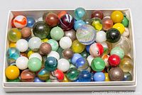 Top view of a white box filled with about 60 mixed antique glass marbles of various colors and designs.