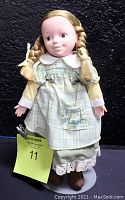 Front view of the Laura Ingalls doll wearing a yellow and green plaid dress with apron embroidered 'Laura', standing on a doll stand against a black background.