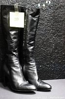Front view of the pair of black knee high leather boots standing upright against a black textured background showing full length and shape.