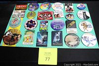 Full layout of 24 assorted button pins on turquoise background with yellow and blue paperwork in front. Pins feature various characters and themes mainly Disney.