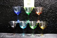 Front view of 7 vintage stemmed champagne coupe martini glasses all different colors, arranged in a display with 3 glasses on top and 4 on the bottom on a black background.