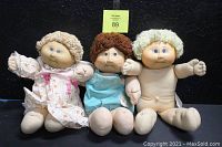 Photo of three Cabbage Patch dolls: one nude with blonde hair, one in white floral dress, one in turquoise onesy. All sitting side by side