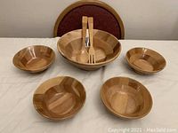 Photo showing one large wooden salad bowl, four smaller wooden bowls, and a pair of wooden serving utensils standing inside the large bowl.
