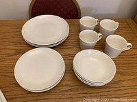 Full set of white dinnerware including three plates, three salad plates, four soup bowls, and four coffee mugs arranged on a wooden table.