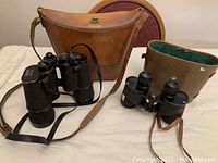 Two pairs of vintage binoculars with two leather carrying cases on a white surface.