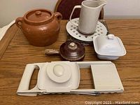 Photo showing a collection of miscellaneous kitchen items on a wooden table including ceramic pots, a pitcher, a butter dish, a vegetable slicer, and a cork stopper.