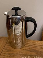Front view of the stainless steel coffee maker showing black plastic handle and lid knob, reflective surface with some wear.